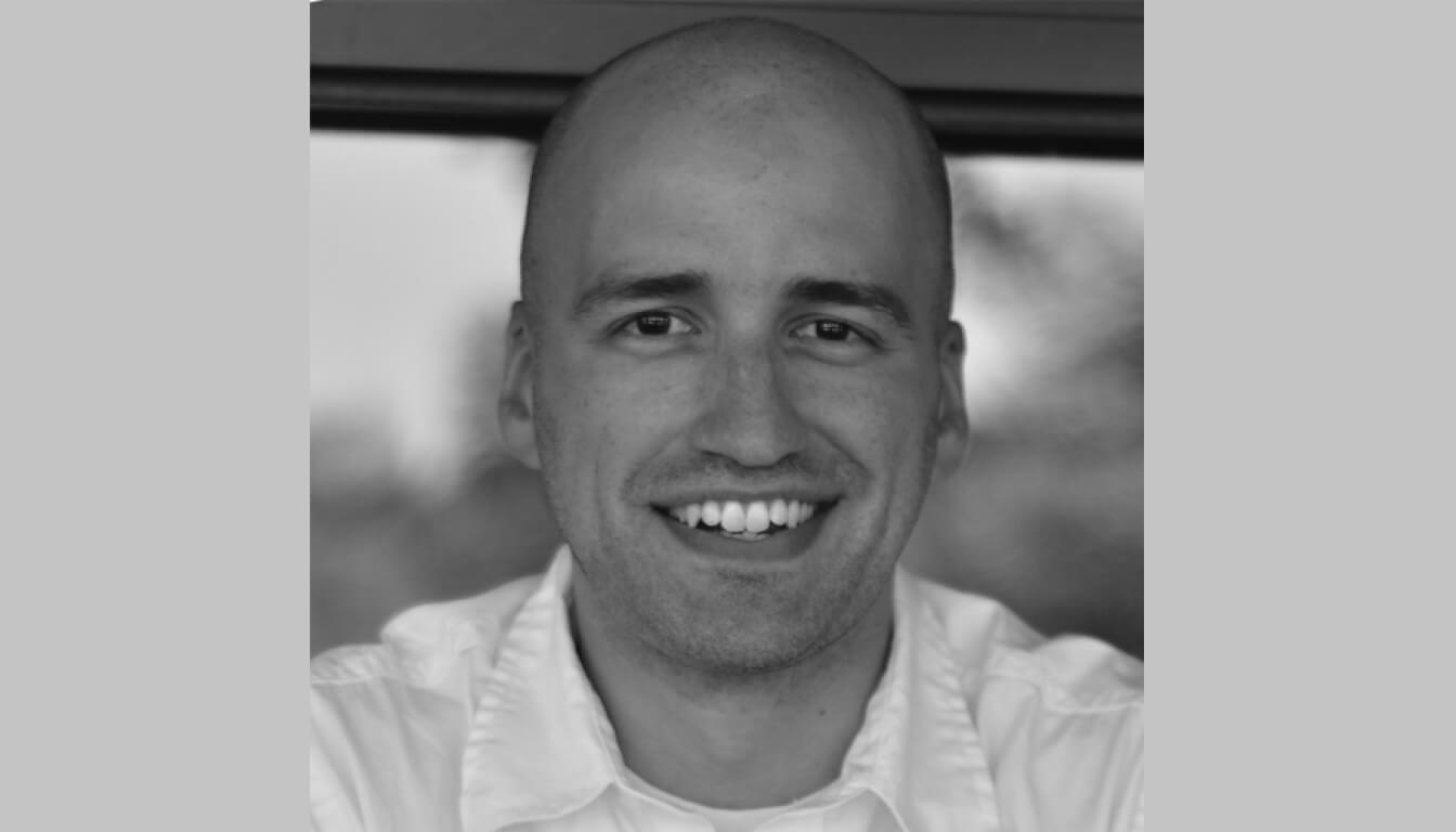 A person with a shaved head grinning warmly, dressed in a collared shirt and captured in an elegant black-and-white portrait.