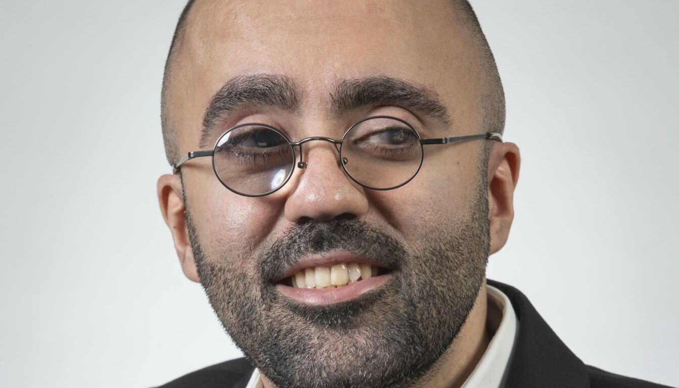 A bearded individual with round glasses and a slight smile looks to the side, dressed in a dapper suit jacket and crisp white shirt.