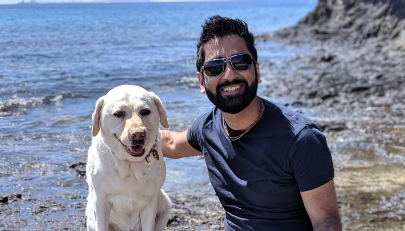 A man in sunglasses and a dark t-shirt smiles while sitting on rocky terrain by the sea, with his loyal white Labrador retriever beside him, basking in the sun.