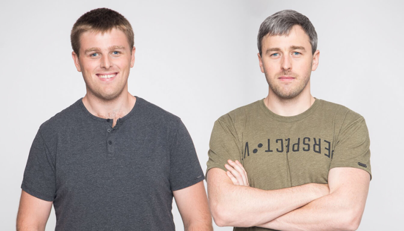 Two men stand against a plain background. The man on the left, smiling, wears a dark T-shirt, while the man on the right has his arms crossed and dons a green T-shirt with text.