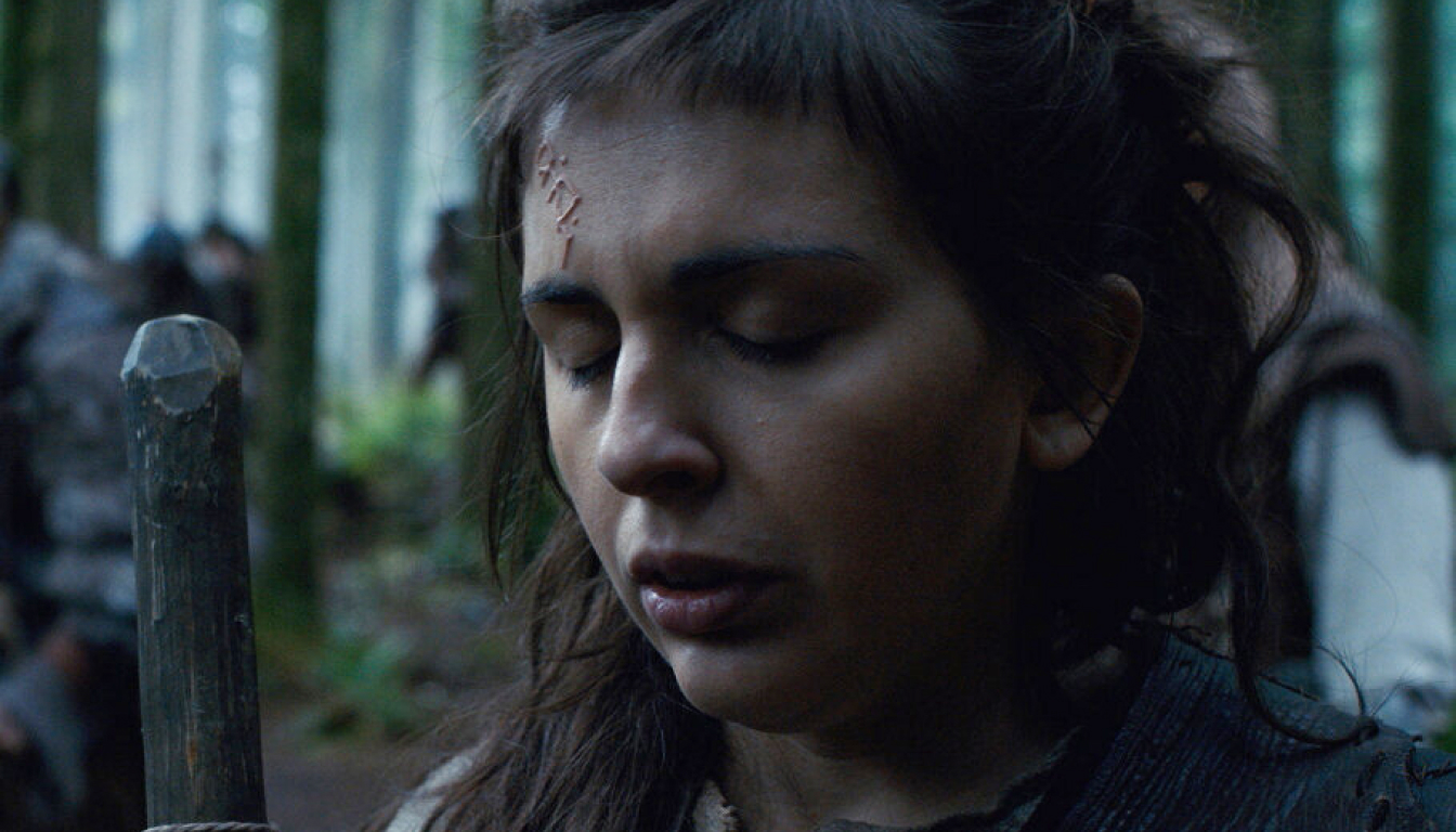 In a mystical forest, a woman with closed eyes and a distinctive scar on her forehead stands solemnly holding an intricately carved staff.