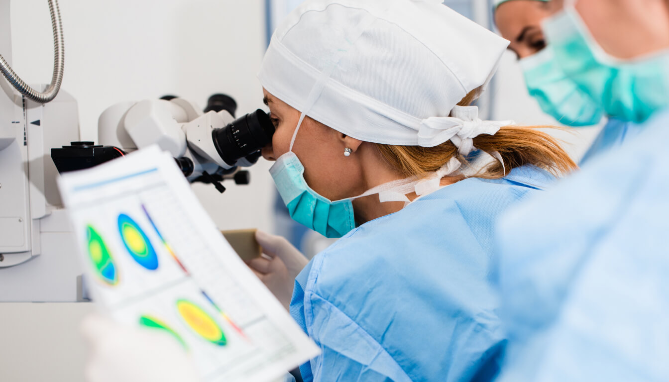 A scientist in blue scrubs and a mask meticulously examines a specimen through a microscope while jotting observations on a chart filled with colorful data.
