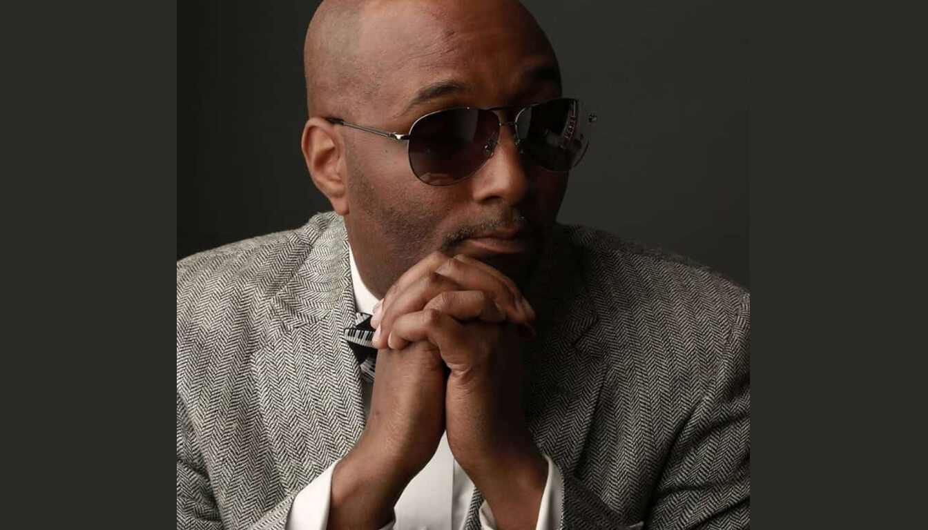 A bald man exudes confidence as he sits against a dark background, wearing sunglasses and a gray herringbone suit, with his hands clasped thoughtfully under his chin.