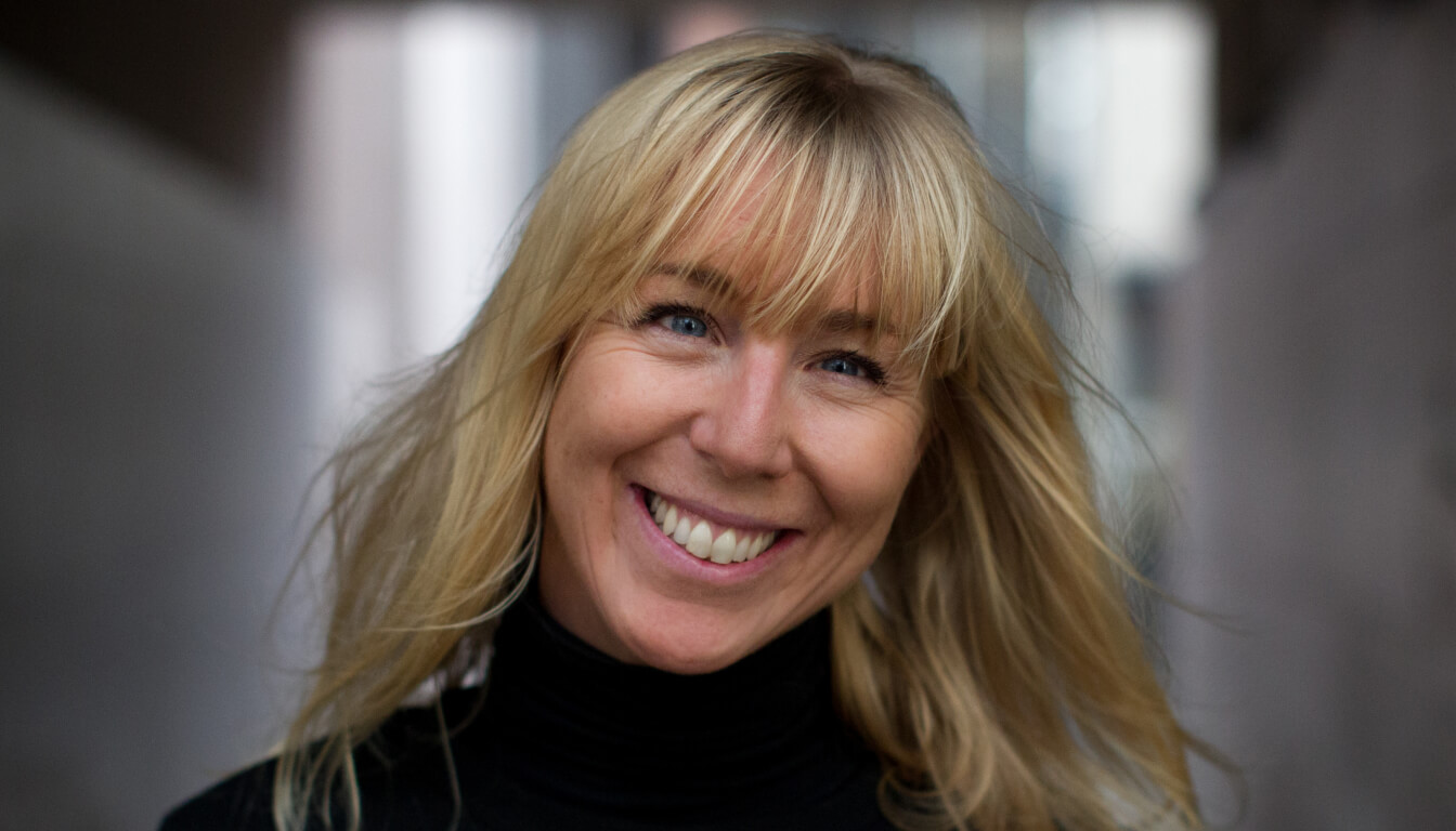 A person with long blond hair and a bright smile is wearing a black turtleneck sweater against a softly blurred background.