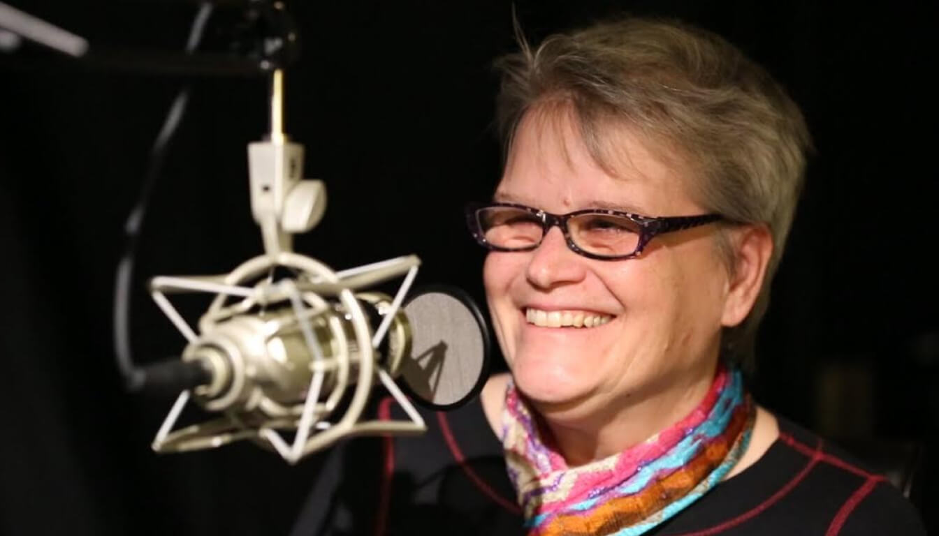 The individual with short hair and glasses is smiling near a microphone, donning a vibrant scarf and dark attire, set against an enigmatic backdrop.