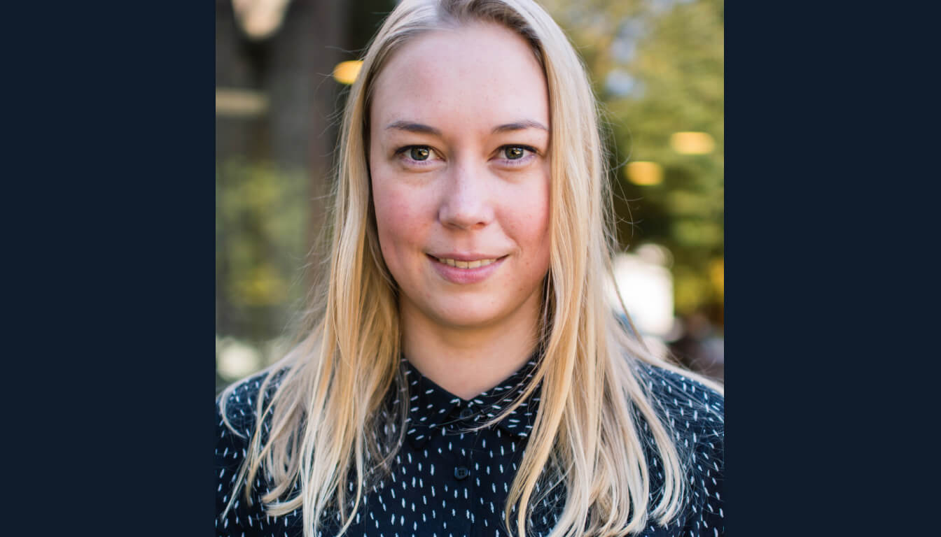 A smiling person with long blonde hair wears a black and white patterned shirt, standing outdoors surrounded by trees under a sunny sky.