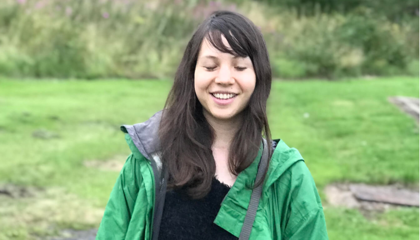 Person smiling with closed eyes, wearing a green jacket, standing outdoors with grassy background.