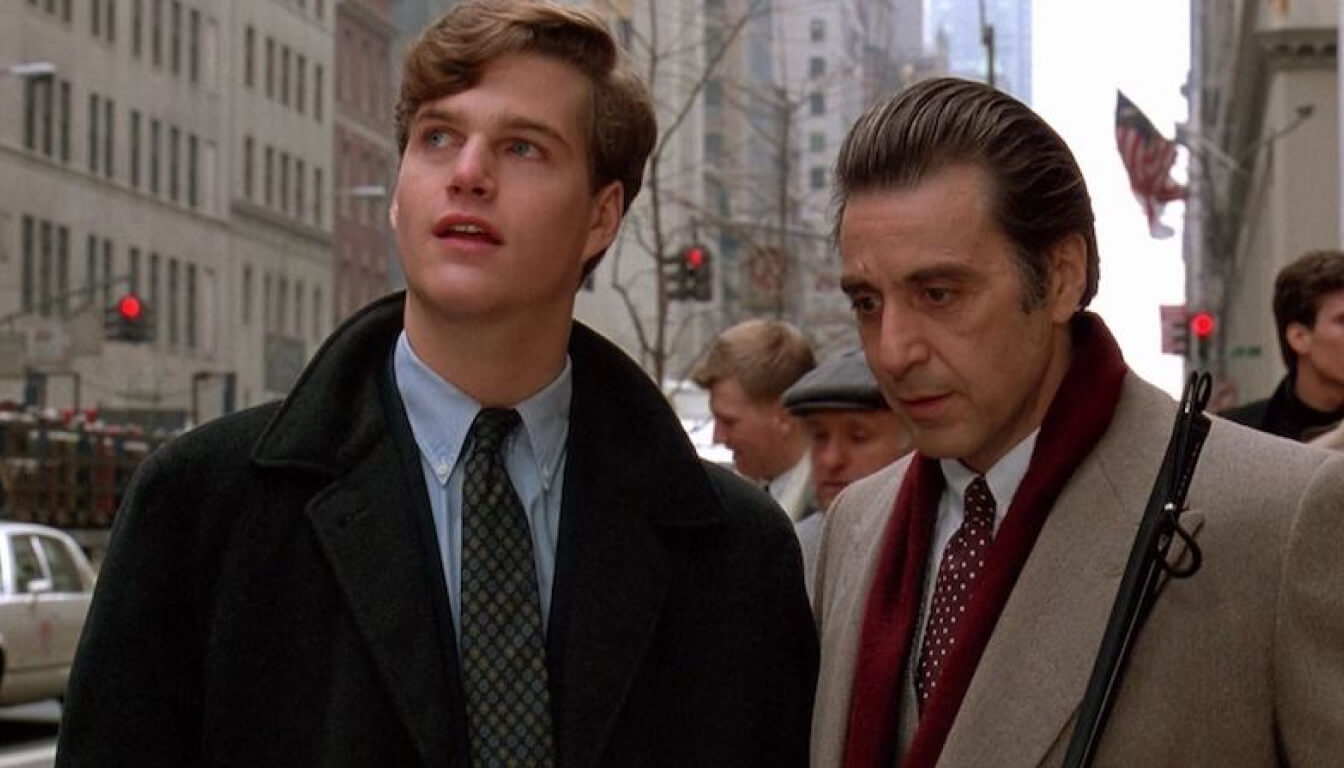 Two men in formal attire stand on a bustling city sidewalk. One, with brown hair, gazes upward, while the older man with dark hair leans on his walking stick. They are surrounded by towering buildings and a throng of pedestrians.