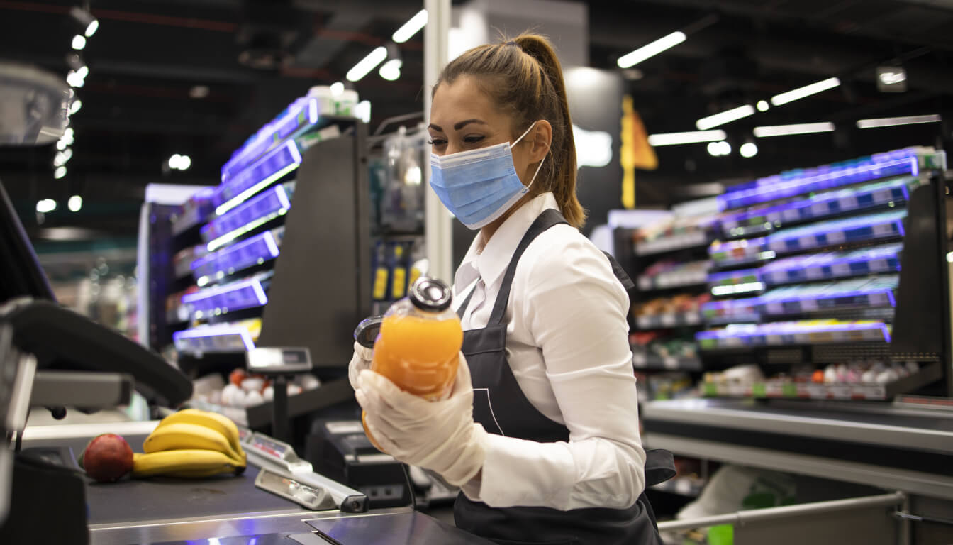 At a bustling supermarket checkout, a cashier wearing a mask and gloves expertly scans a bottle of fresh orange juice.