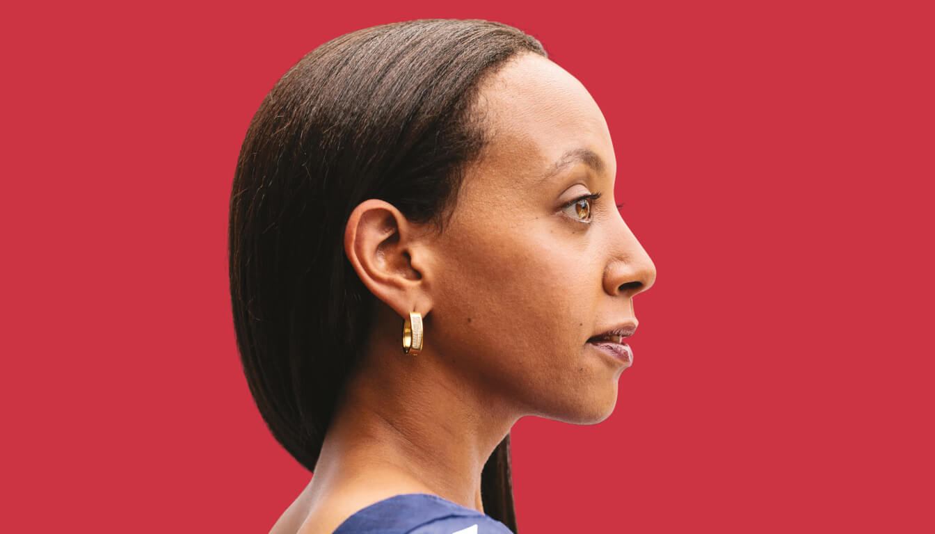 Profile view of a person with long dark hair, wearing gold hoop earrings and a blue top, set against a vivid red background.