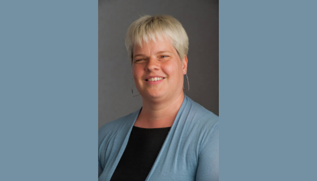 A smiling person with short blonde hair wears a light blue cardigan over a black top, standing against a neutral background, exuding warmth and confidence.