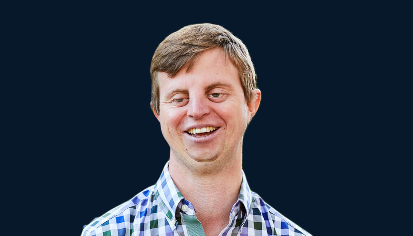 A person with a friendly expression, wearing a checkered shirt and exuding confidence, is shown against a solid dark background.