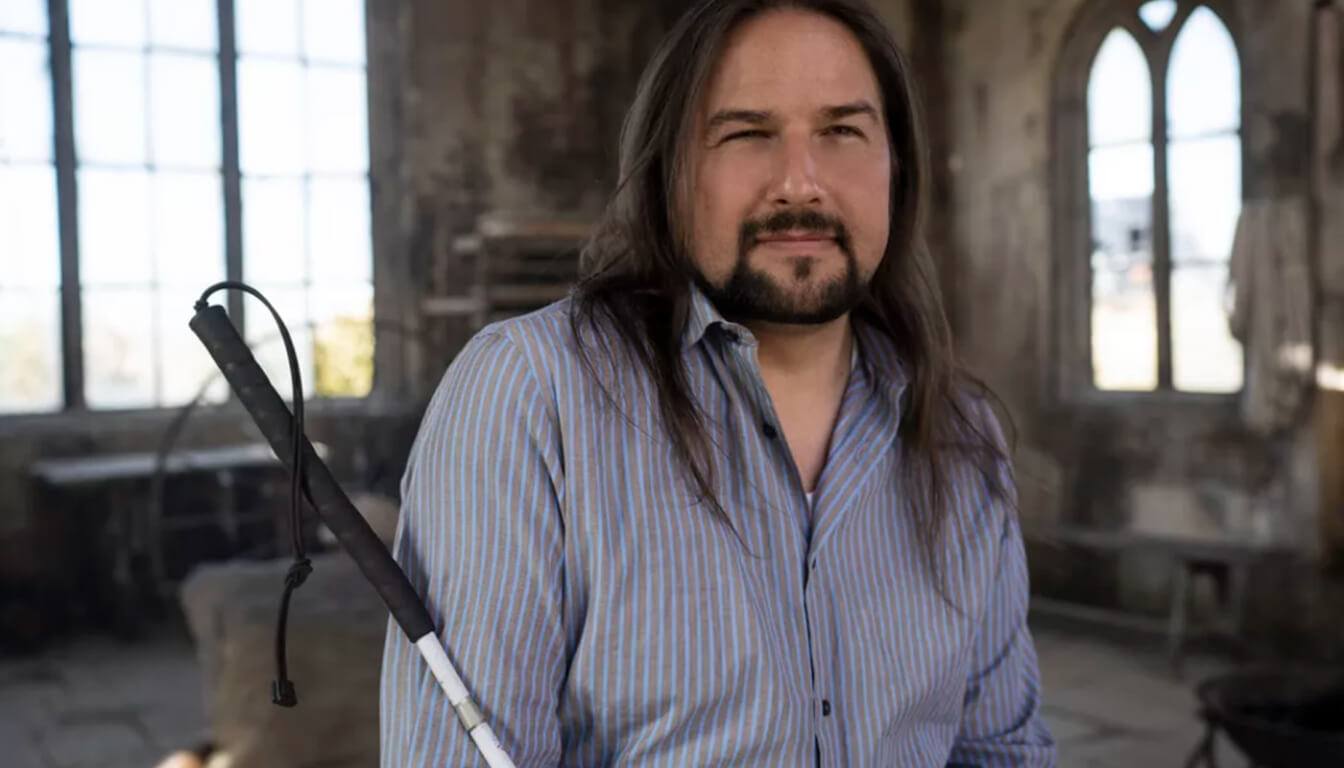 A man with long hair stands in a rustic room, gripping a white cane. He's clad in a striped button-up shirt, while large windows and worn walls provide the backdrop. The sunlight streaming through gives the space an inviting, nostalgic charm.