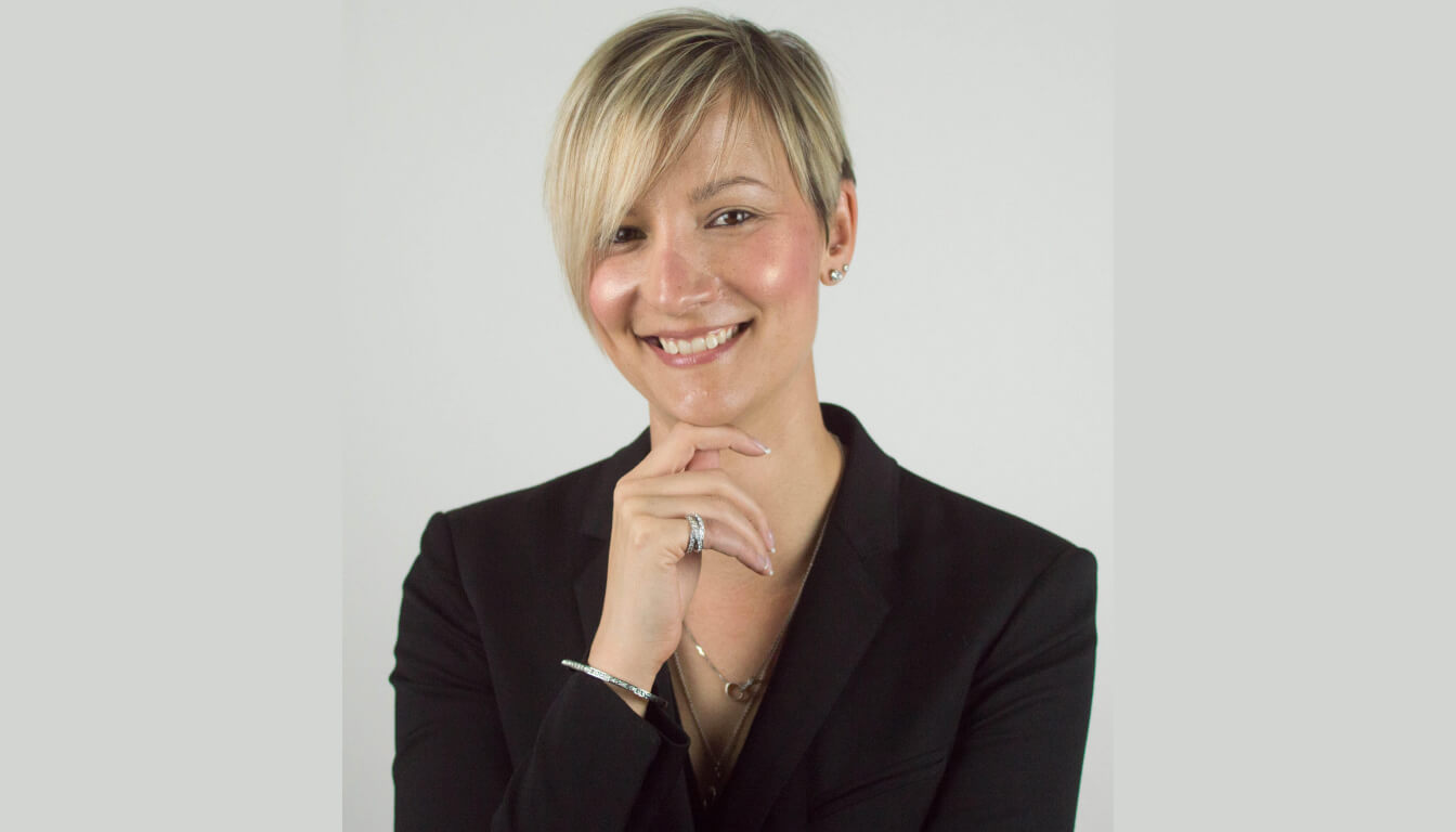 A person with short blond hair is smiling and wearing a black blazer, exuding confidence. Their hand rests thoughtfully under their chin against a plain, light-colored background.