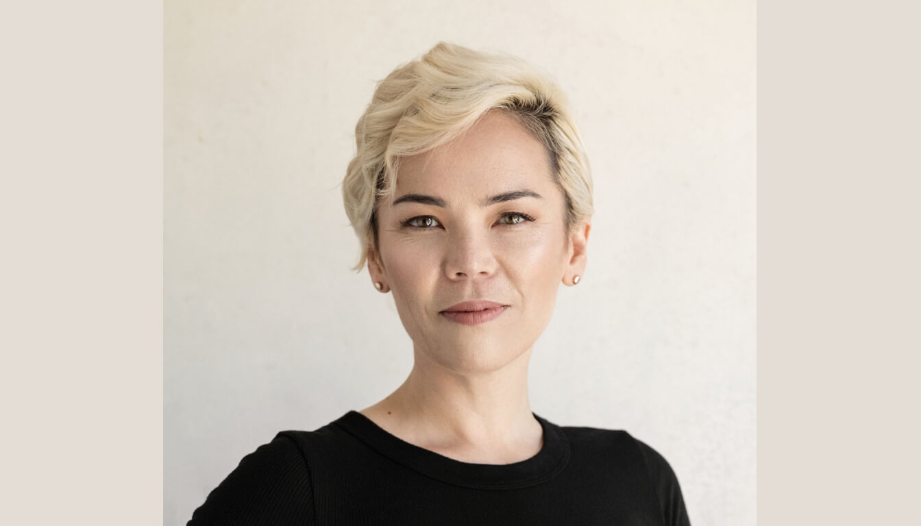 A person with short blonde hair and a black shirt gazes directly at the camera, set against a minimalist backdrop.