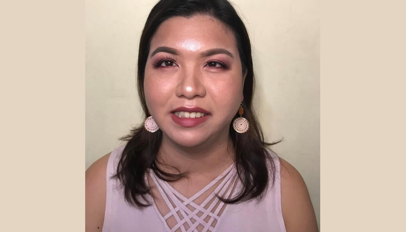 A woman with long dark hair and pink eyeshadow smiles, showcasing her patterned earrings. She dons a light-colored top with a lattice design, exuding elegance.