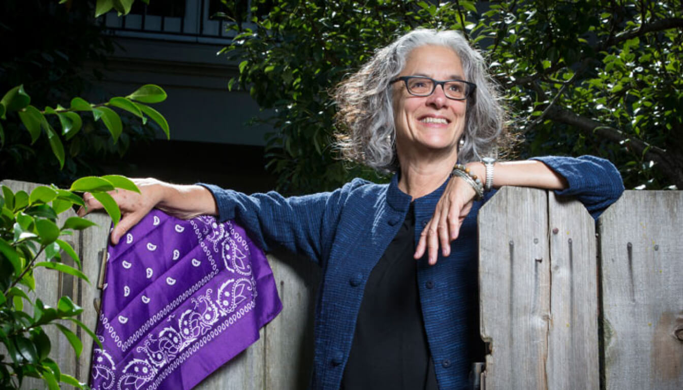 A person with gray hair and glasses, clad in a blue jacket, leans on a wooden fence adorned with a purple bandana, while lush trees create a serene backdrop.