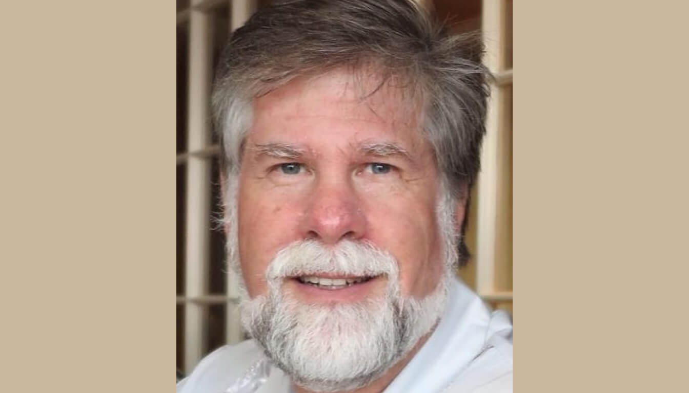 A man with gray hair and a beard is smiling warmly at the camera. He is dressed in a light-colored shirt, set against a softly blurred background that reveals window panels and sunlight filtering through.