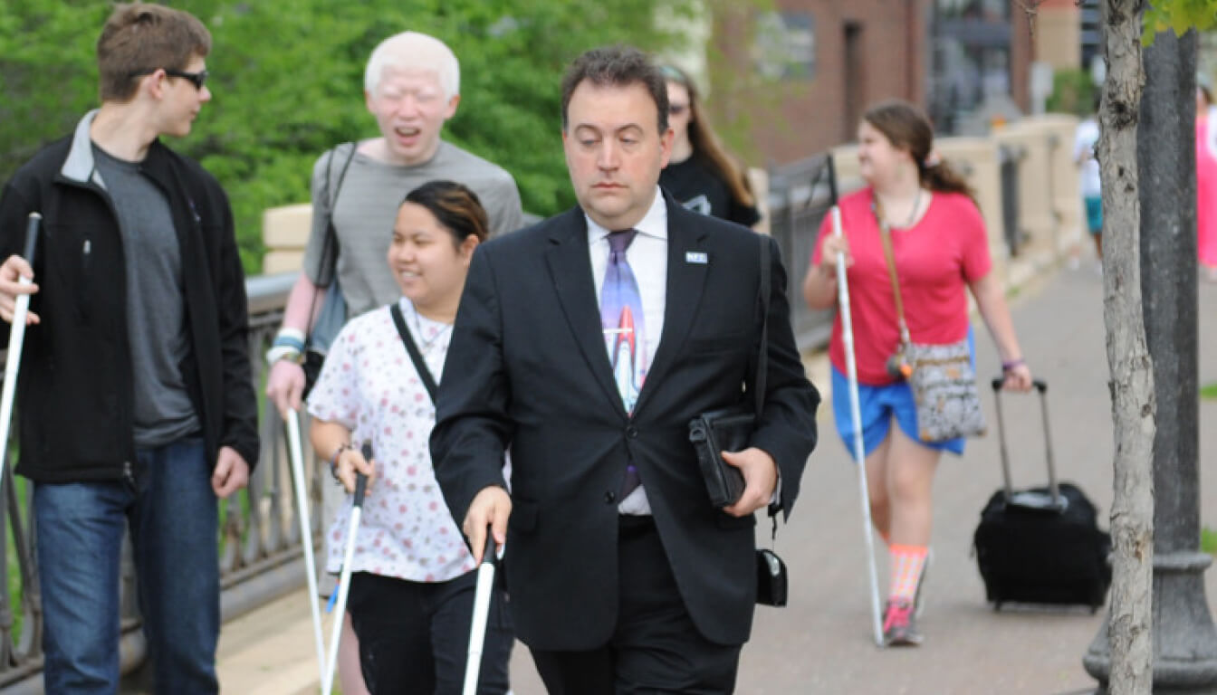 A group moves together on the sidewalk, one man in a suit leading and others casually dressed. Among them are individuals with walking canes. Trees and a building serve as the backdrop, creating a lively urban scene.