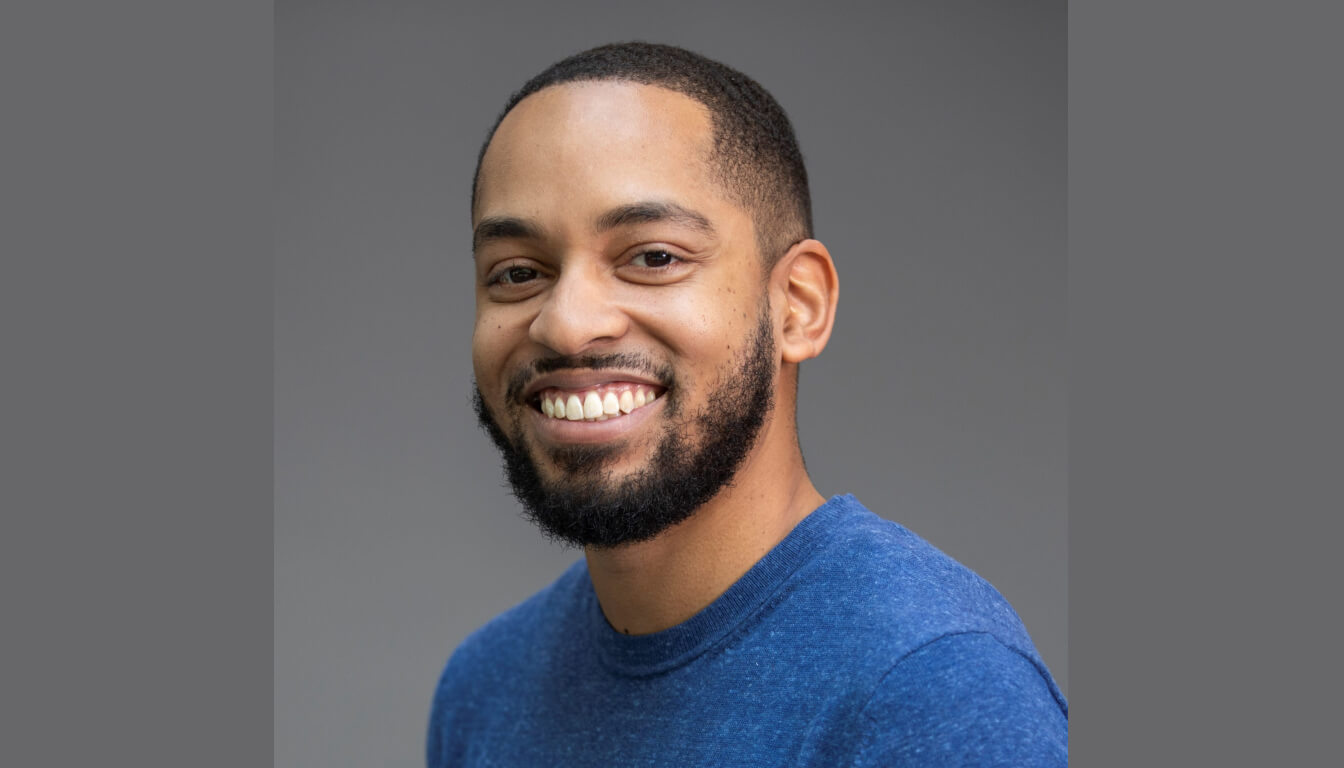 A cheerful man with a short haircut and a beard stands out in a blue shirt against a gray backdrop.