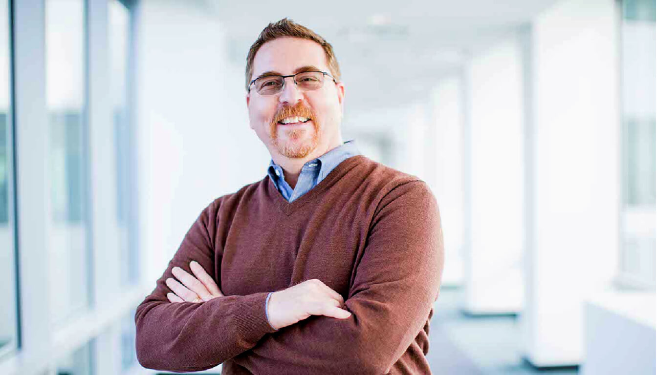A man with glasses and a well-groomed beard stands confidently in a hallway, arms crossed, wearing a cozy brown sweater over a crisp blue collared shirt.