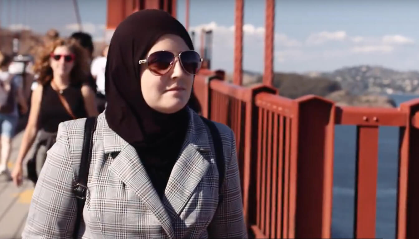 A woman confidently strides across a bridge, her sunglasses gleaming under the sun. Her hijab billows gently in the breeze as others walk behind her, painting a lively scene of movement and style.