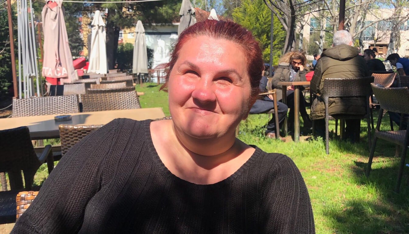 A person with red hair in a black sweater enjoys a sunny day at a café. Umbrellas and tables can be seen in the background, creating a lively atmosphere.