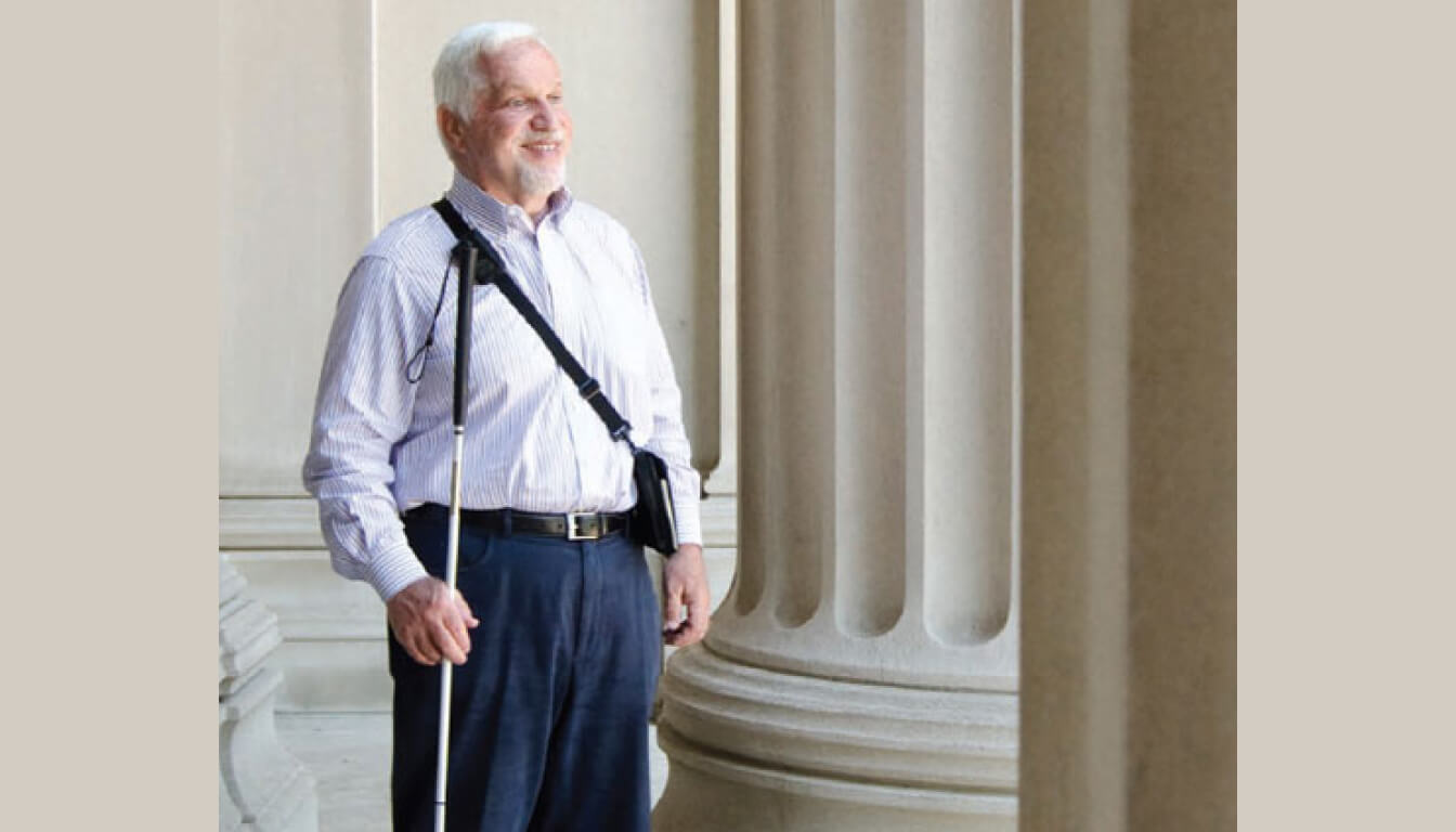 A man stands by large columns, wearing a striped shirt and dark pants, with a strap over his shoulder. Holding a white cane, he looks ahead and smiles confidently.