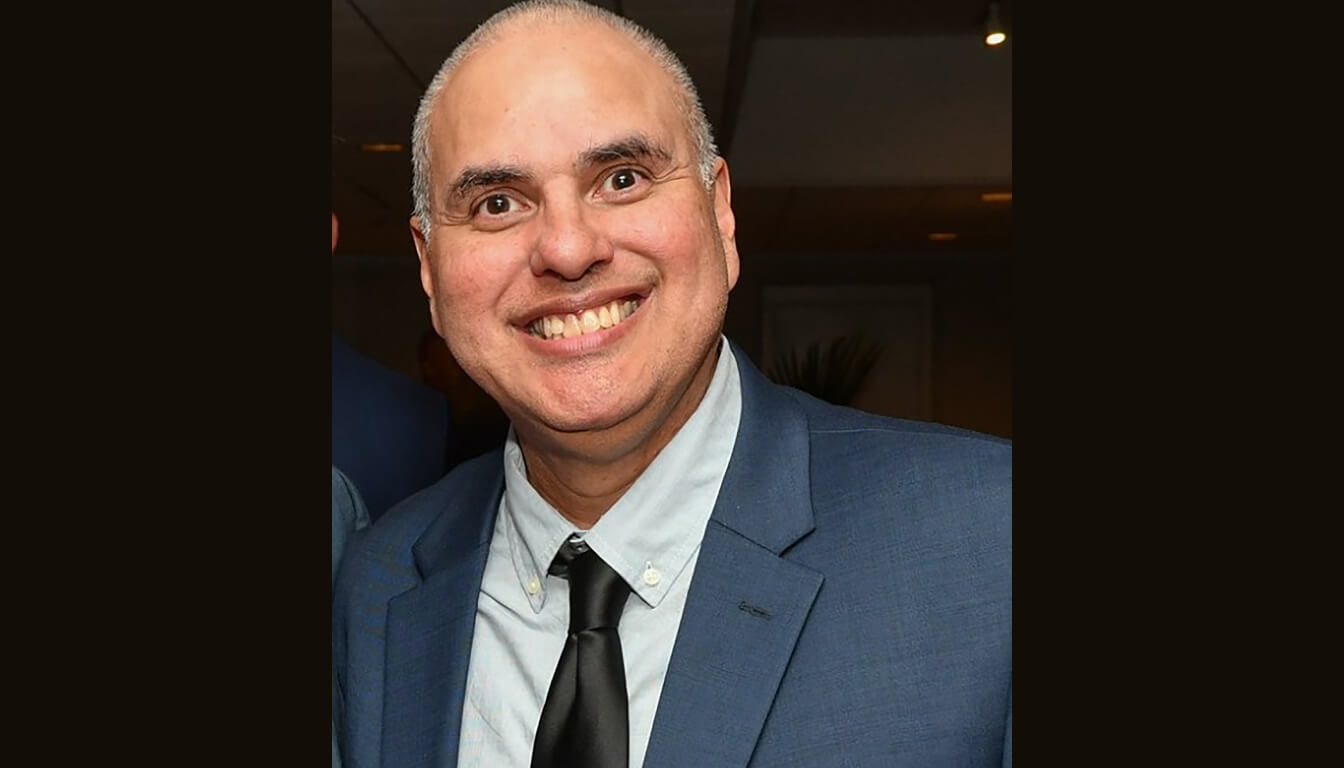 A cheerful man in a blue suit and black tie stands confidently against a dark backdrop, exuding elegance and charm.