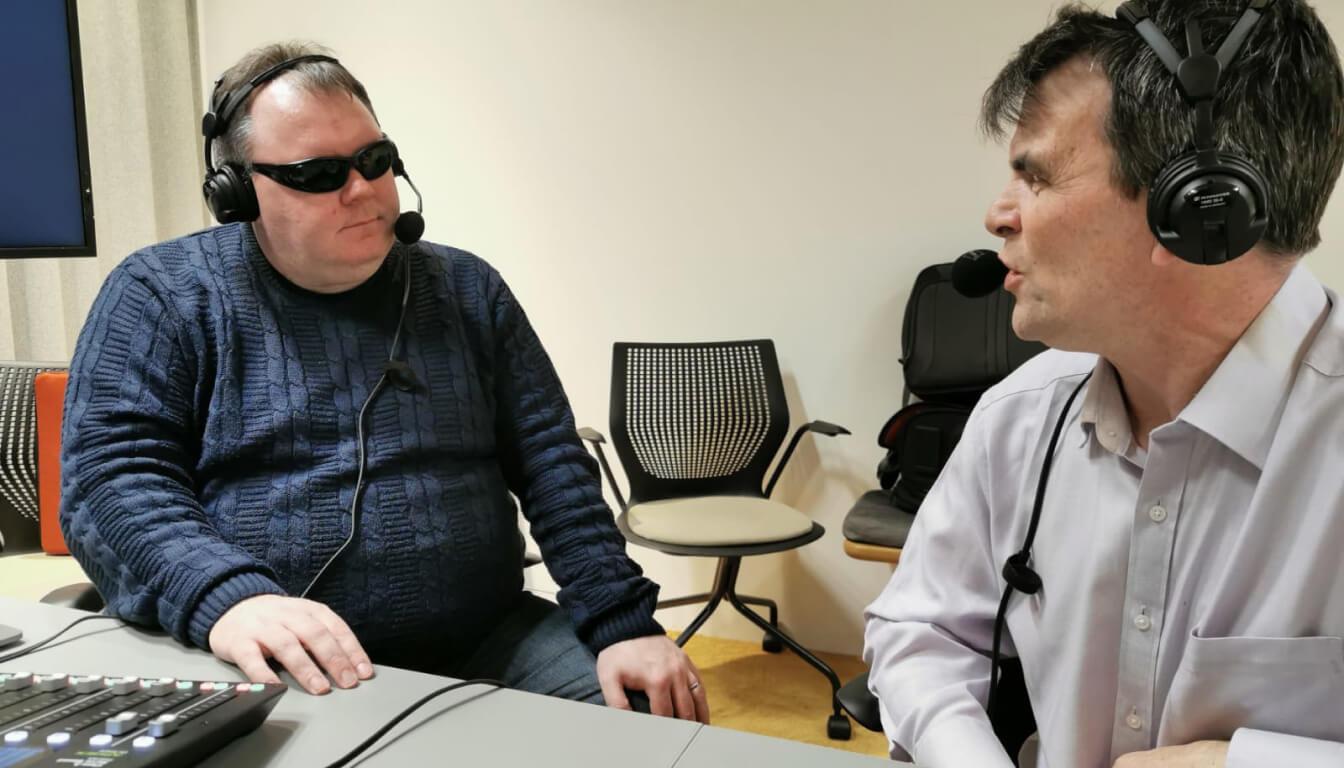 Two men wearing headsets engage in conversation at a table with audio equipment. One sports sunglasses and a blue sweater, while the other wears a light shirt. Empty chairs linger in the background, hinting at a lively discussion or an upcoming recording session.