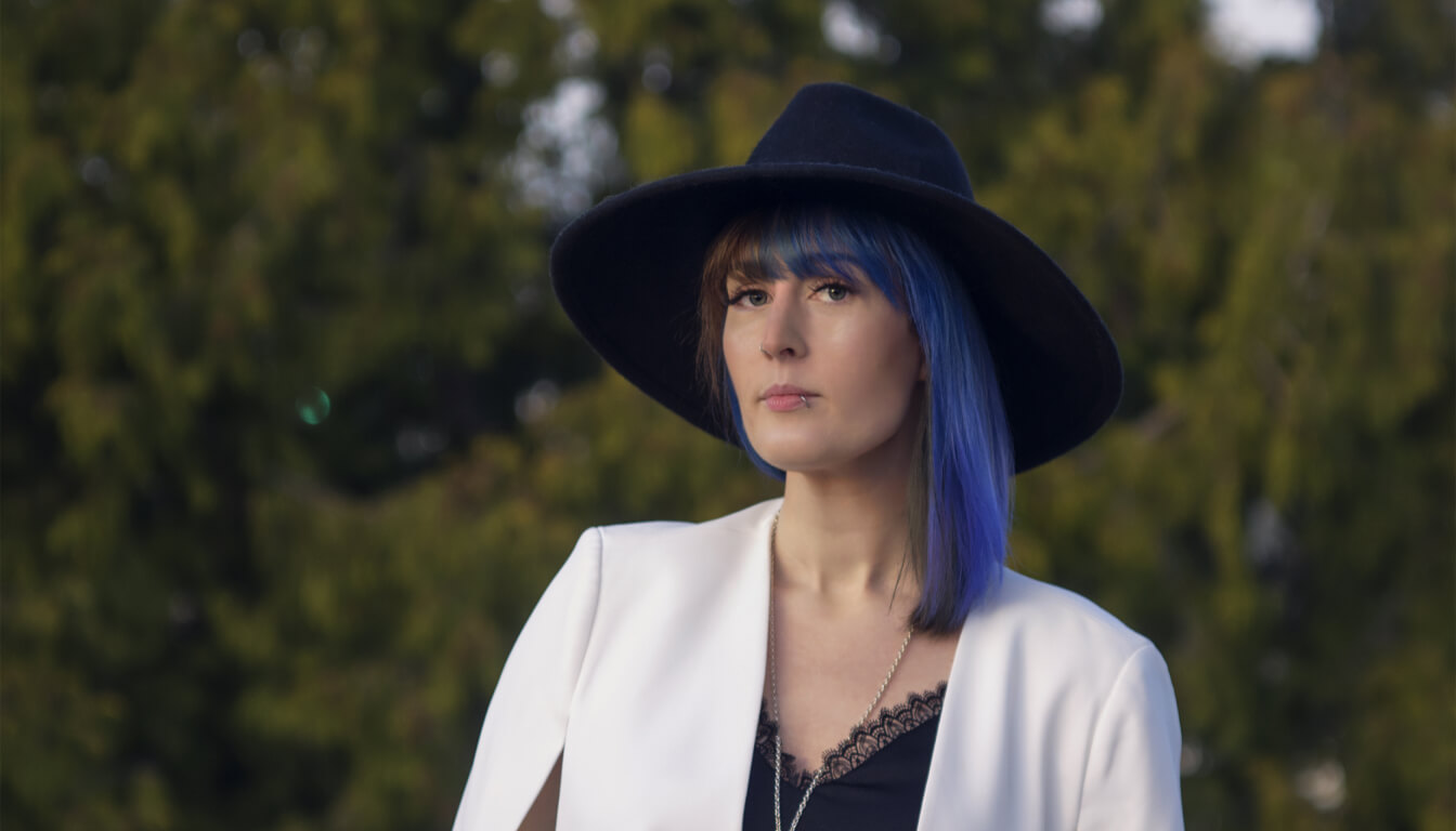 A person with striking blue hair is wearing a black wide-brimmed hat, a white blazer, and a black top. They stand outdoors against the backdrop of lush trees, exuding an air of style and confidence.