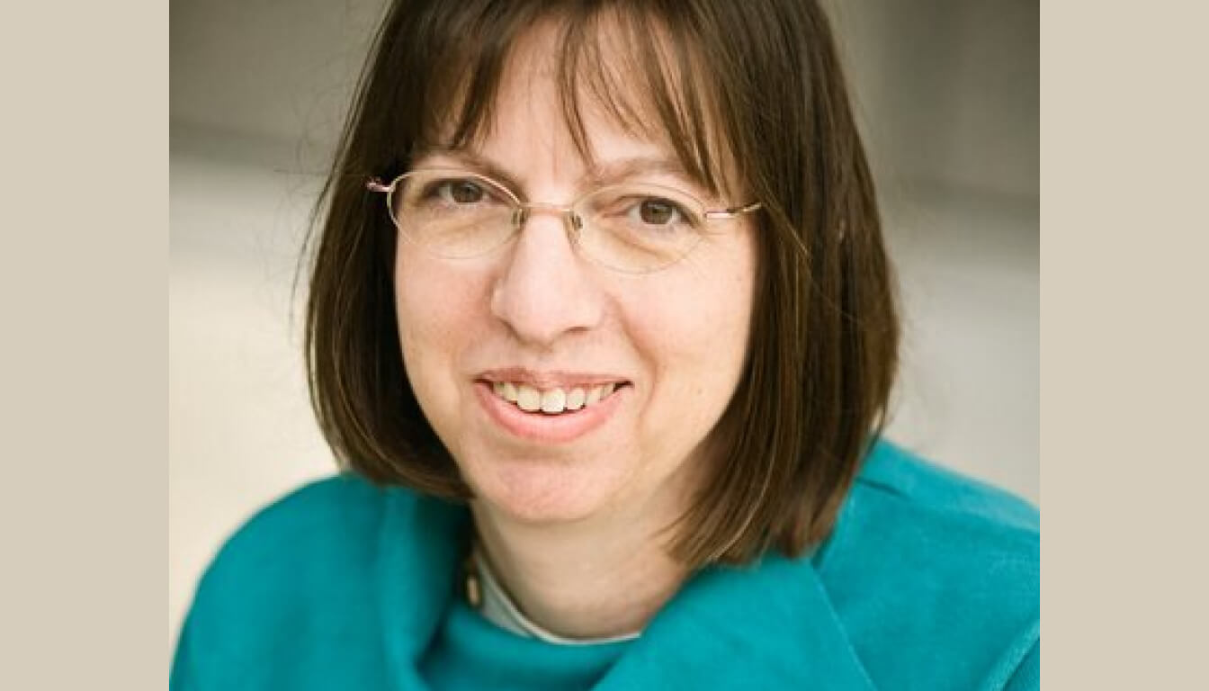 A person with shoulder-length brown hair and glasses is smiling brightly, wearing a stylish teal jacket that accentuates their cheerful demeanor.