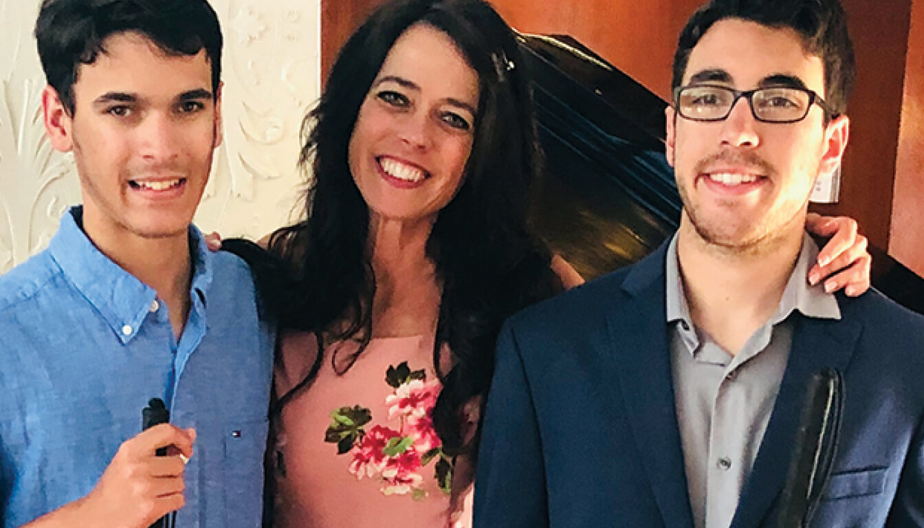 Three people are posing and smiling indoors. A woman in a floral dress stands between two young men, one wearing a blue shirt and the other dressed in a suit.