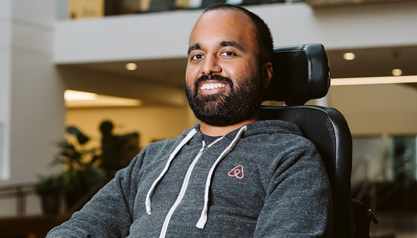 A person with a beard and a friendly smile sits in a wheelchair, leisurely wearing a cozy gray hoodie indoors. The room is filled with warmth and positivity.