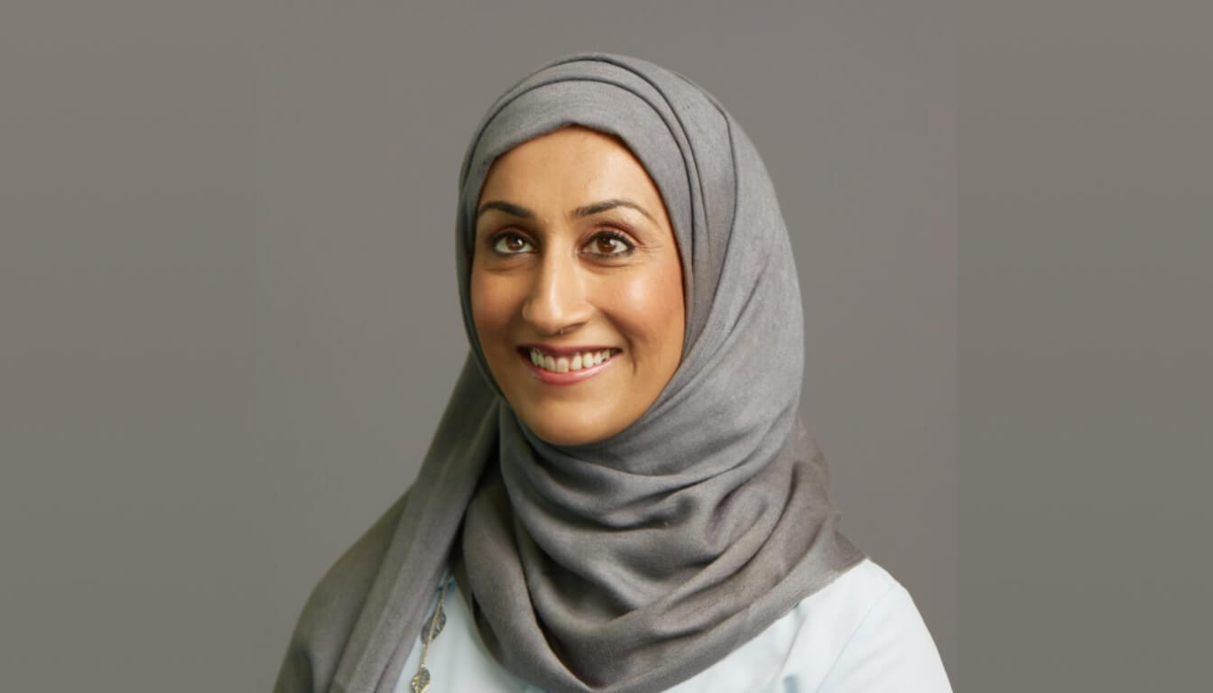 A cheerful individual in a gray hijab and light top beams with a warm smile against a simple backdrop.