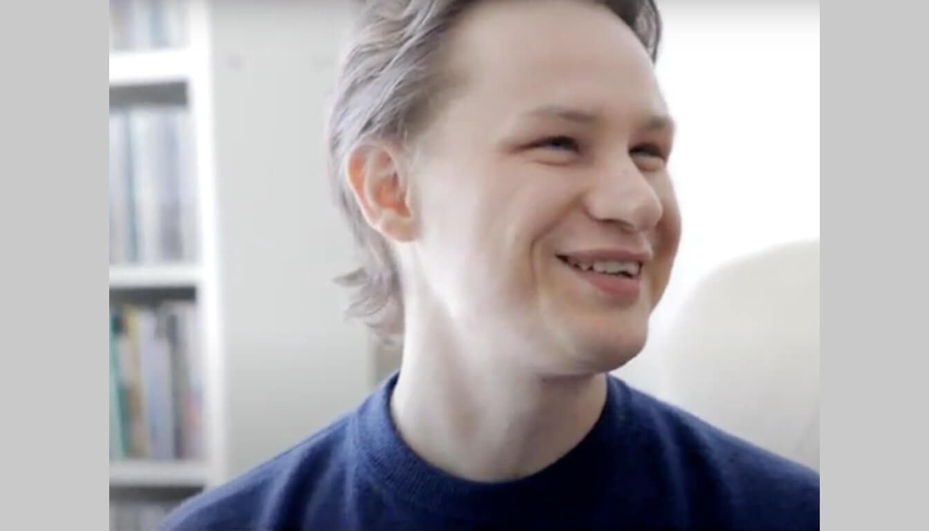 Person with short hair smiling warmly, wearing a dark sweater, and standing before a softly blurred bookshelf.