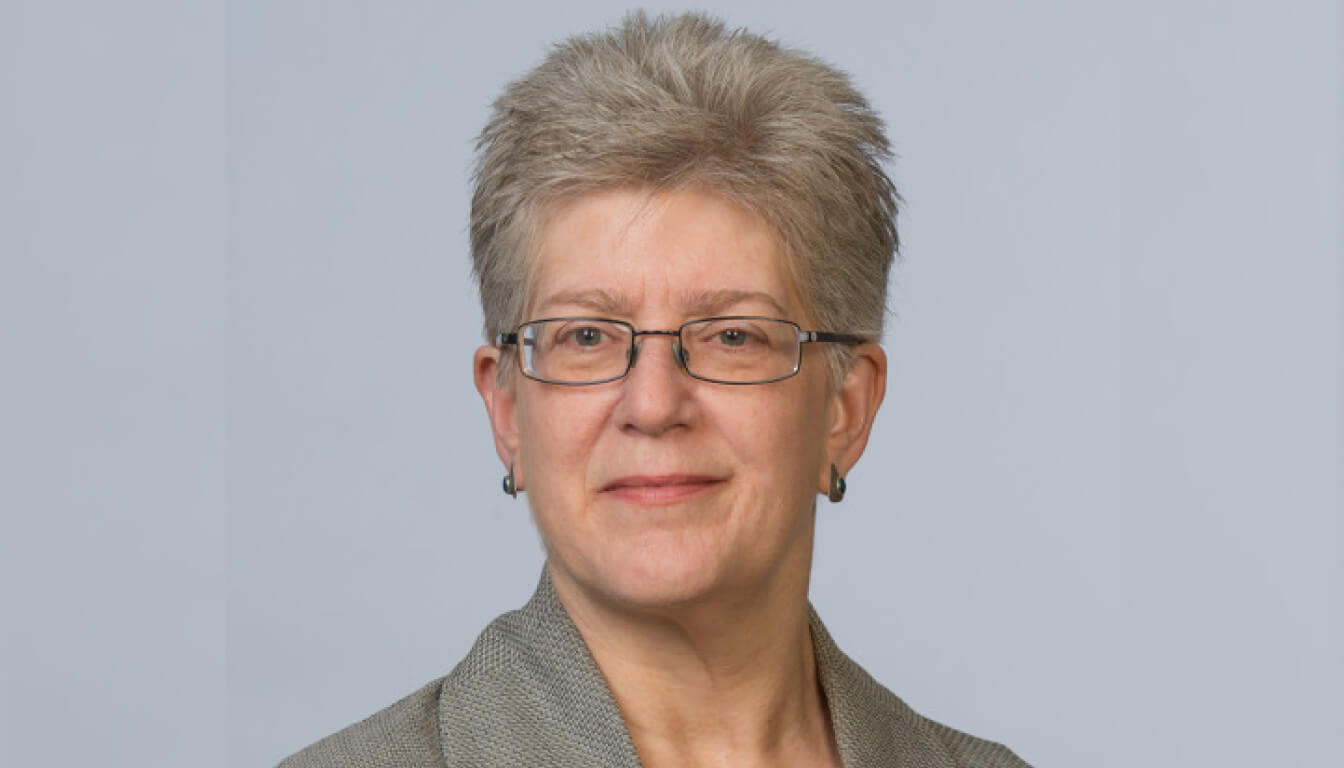 A distinguished individual with short gray hair and glasses, dressed in a sophisticated gray blazer, posed against a neutral backdrop.