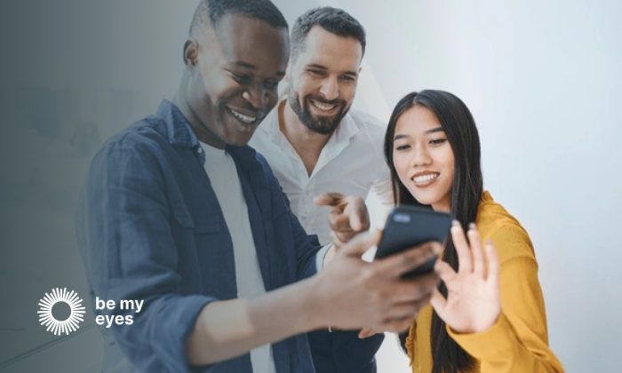 Three people smiling and looking at a smartphone together. One person is holding the phone. The "Be My Eyes" logo is in the bottom left corner.