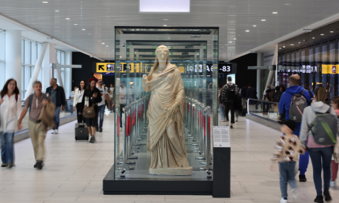 Marble statue of a draped figure in a glass case, displayed in a busy airport terminal with travelers walking by.