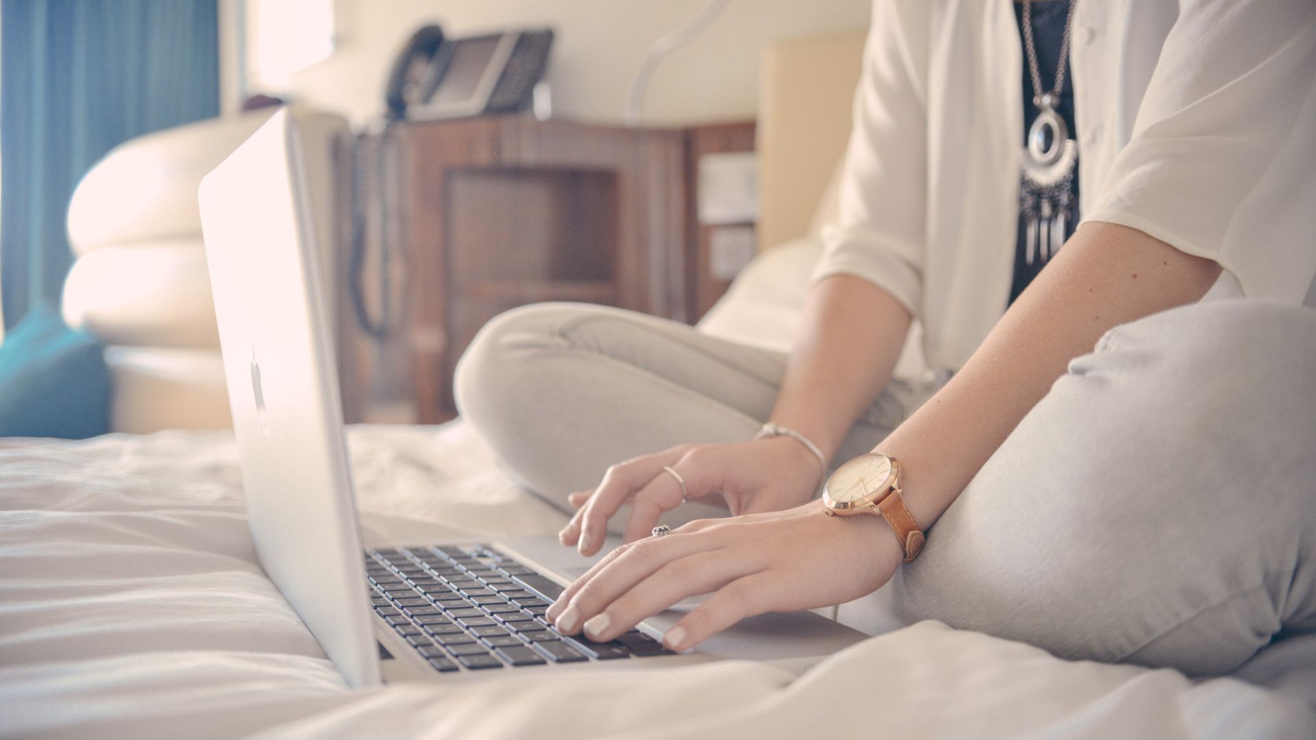Person sitting cross-legged on a bed, typing on a laptop, wearing light-colored clothes and a watch, with a blurred background showing furniture.