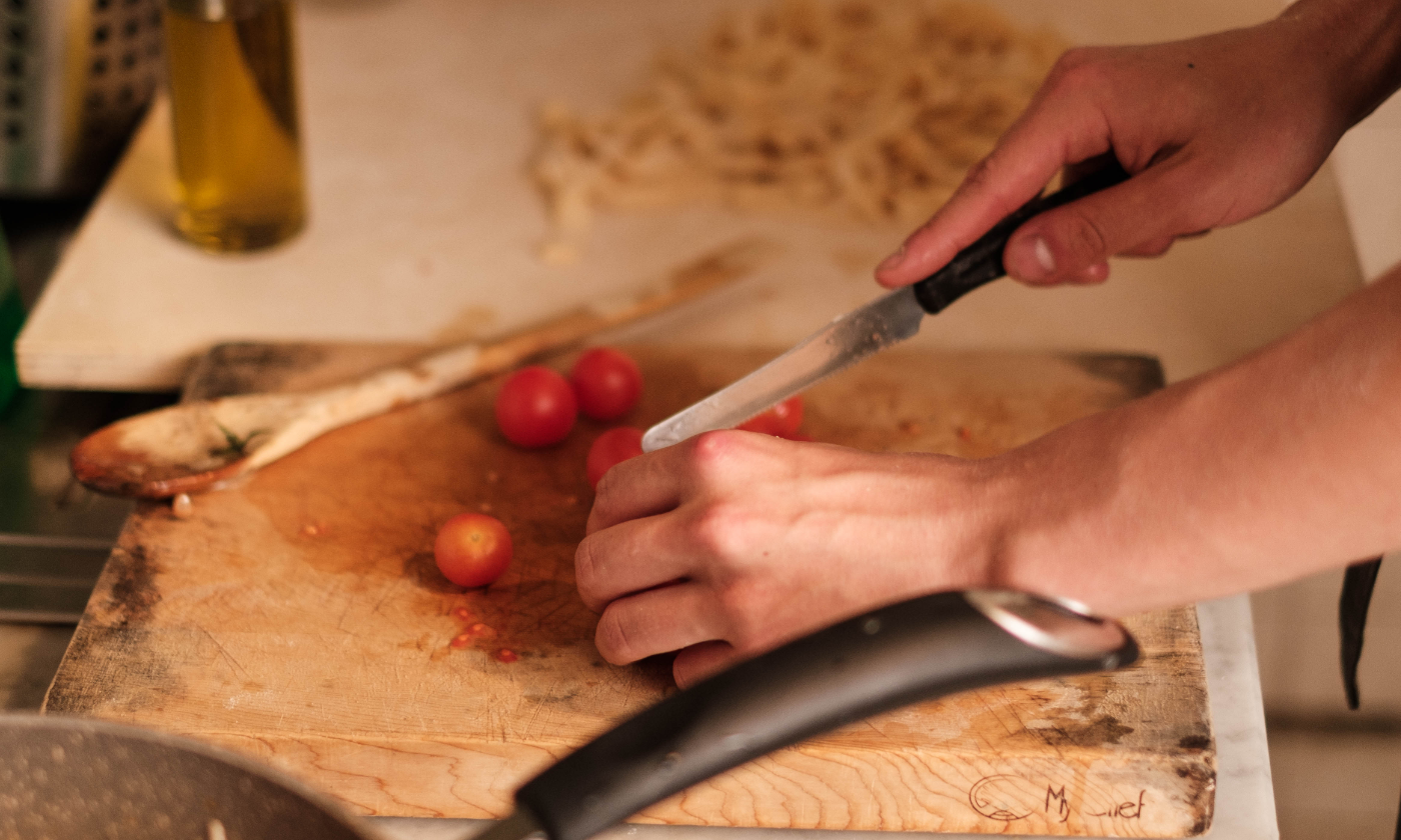 A person slicing cherry tomatoes on a wooden cutting board with a knife, surrounded by kitchen utensils and ingredients.