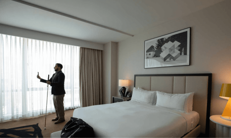 A man holding a white cane stands by the window in a hotel room with a neatly made bed, a large window, and modern decor.