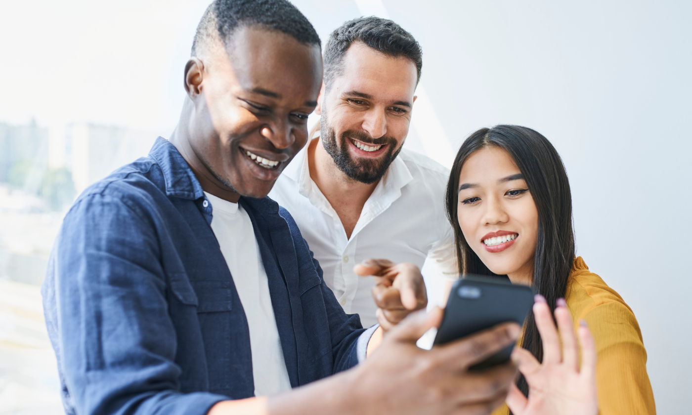 Three people stand together, smiling and looking at a smartphone as one of them holds it up for the group to see.