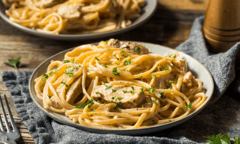 A plate of creamy chicken alfredo pasta garnished with chopped parsley, set on a wooden table with a fork and napkin nearby.